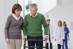 Wife helping husband with walker. Doctors and physical therapist are standing in the background.